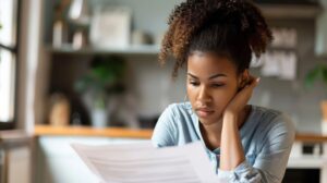 Desapropriação: a imagem mostra uma mulher jovem e concentrada analisando documentos em um ambiente doméstico. Ela parece estar absorvida no conteúdo, com expressão séria e levemente preocupada, o que transmite a ideia de reflexão e tomada de decisões importantes. O fundo desfocado sugere que está em sua casa ou em um local informal, reforçando a sensação de introspecção. Essa cena é ideal para ilustrar temas como despropriação, processos legais ou decisões críticas que envolvem propriedade ou direitos pessoais.