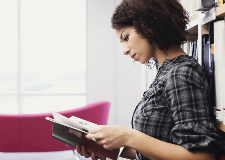 A imagem mostra uma jovem em pé, concentrada enquanto lê um livro em um ambiente iluminado e tranquilo, possivelmente uma biblioteca ou sala de leitura. Ela está ao lado de uma estante com livros organizados, vestindo uma camisa xadrez casual. A composição destaca seu foco e interesse no material, enquanto o fundo desfocado apresenta uma poltrona rosa e uma janela com luz natural entrando no espaço. Essa cena é ideal para abordar temas como estudo, pesquisa, aprendizado ou tomada de decisões informadas, especialmente em contextos de planejamento, análise de direitos ou questões legais, como despropriação.