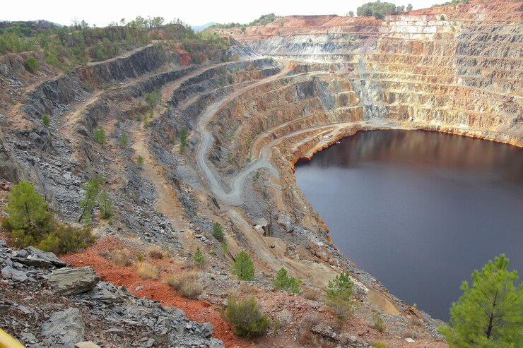 Instalação de Minas: Fotografia de uma mina a céu aberto com camadas em formato de terraços, ao redor de um grande lago formado no fundo da escavação. A paisagem inclui áreas rochosas expostas e vegetação esparsa ao redor, destacando os impactos ambientais da mineração, como a remoção de solo e mudanças na geografia natural. Ideal para ilustrar artigos sobre os impactos ambientais e sociais da instalação de minas.