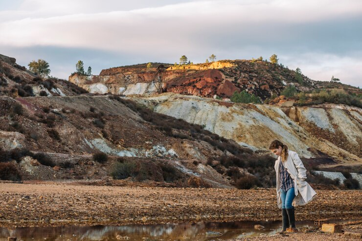 Direitos do superficiário. Instalação de minas: A imagem mostra uma paisagem de exploração mineral, com relevos desgastados e solo exposto, característico de áreas mineradas. No centro, uma mulher caminha próximo a uma área de água superficial, vestida com botas e um casaco, sugerindo que está analisando ou inspecionando o local.