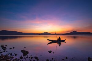 Rompimento de barragens Esta imagem retrata um cenário tranquilo de um pescador solitário em um barco, navegando em um lago durante o pôr do sol. O céu apresenta tons vibrantes de azul, laranja e rosa, refletidos suavemente na superfície da água. Montanhas ao fundo adicionam profundidade ao horizonte, enquanto rochas e pedras na margem em primeiro plano criam um contraste natural.