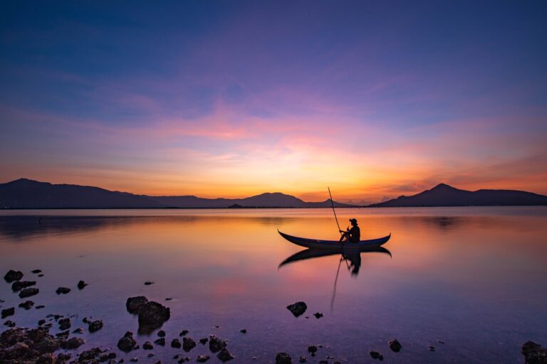 Rompimento de barragens Esta imagem retrata um cenário tranquilo de um pescador solitário em um barco, navegando em um lago durante o pôr do sol. O céu apresenta tons vibrantes de azul, laranja e rosa, refletidos suavemente na superfície da água. Montanhas ao fundo adicionam profundidade ao horizonte, enquanto rochas e pedras na margem em primeiro plano criam um contraste natural.