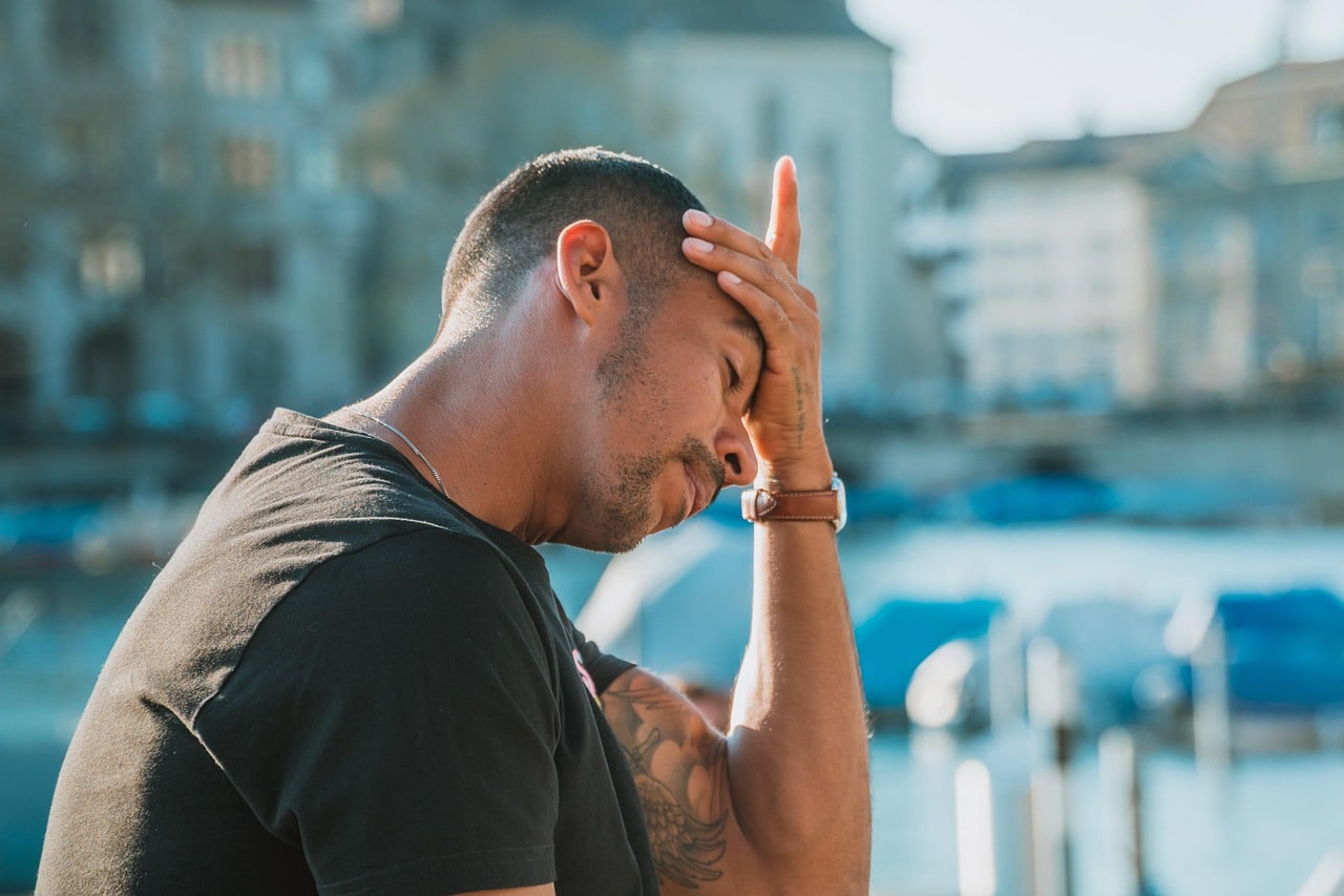 Processo de desapropriação - A imagem mostra um homem com expressão de preocupação, segurando a cabeça com a mão, como se estivesse angustiado. O cenário ao fundo sugere um ambiente urbano próximo à água. A expressão dele pode estar relacionada a problemas financeiros, pessoais ou até mesmo a um processo de desapropriação, que pode ter causado impacto em sua vida.