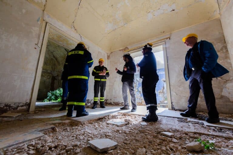 A imagem mostra uma equipe de inspeção em um edifício em ruínas, avaliando sua estrutura. Vestindo uniformes de segurança e capacetes, eles parecem discutir questões técnicas, possivelmente ligadas à **utilidade pública**, como vistorias de segurança ou desapropriação.
