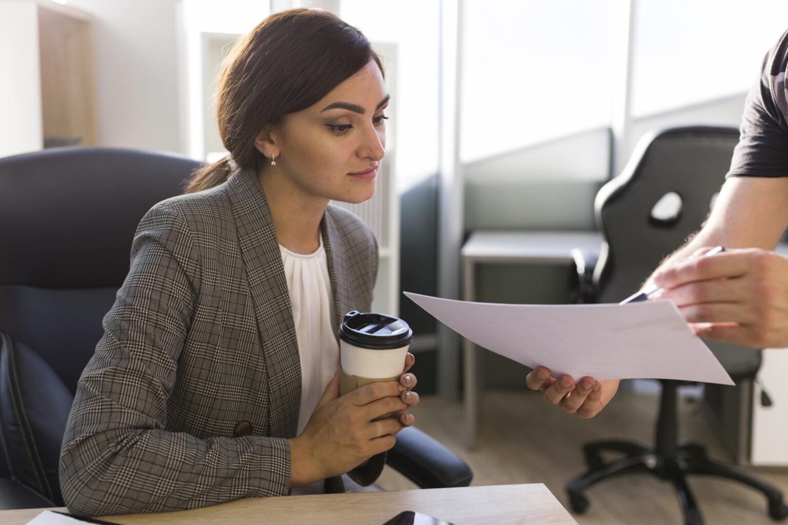 consultoria trabalhista A imagem mostra uma mulher em um ambiente de escritório, com expressão atenta e postura profissional. Ela segura um copo de café enquanto analisa um documento apresentado por outra pessoa, que está parcialmente fora de foco. A cena transmite a ideia de uma reunião ou orientação técnica, representando o momento em que uma especialista em consultoria trabalhista revisa documentos, contratos ou informações jurídicas de um cliente. O ambiente corporativo, a roupa formal e a interação entre os dois profissionais reforçam o conceito de assessoria especializada, voltada para a análise de direitos, deveres e conformidade com a legislação trabalhista.