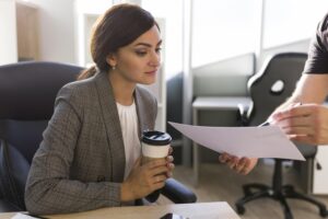 consultoria trabalhista A imagem mostra uma mulher em um ambiente de escritório, com expressão atenta e postura profissional. Ela segura um copo de café enquanto analisa um documento apresentado por outra pessoa, que está parcialmente fora de foco. A cena transmite a ideia de uma reunião ou orientação técnica, representando o momento em que uma especialista em consultoria trabalhista revisa documentos, contratos ou informações jurídicas de um cliente. O ambiente corporativo, a roupa formal e a interação entre os dois profissionais reforçam o conceito de assessoria especializada, voltada para a análise de direitos, deveres e conformidade com a legislação trabalhista.