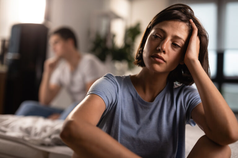 desapropriação: A imagem mostra uma mulher em primeiro plano, sentada, com expressão de tristeza ou preocupação. Ela apoia a cabeça em uma das mãos, parecendo pensativa ou abatida. Ao fundo, desfocado, há uma outra pessoa — possivelmente um homem — também sentada, sugerindo que os dois podem ter tido uma discussão ou estarem passando por um momento de tensão. O ambiente parece ser doméstico, com iluminação suave e um clima introspectivo. A cena transmite sentimentos de distância emocional, reflexão e conflito interpessoal.