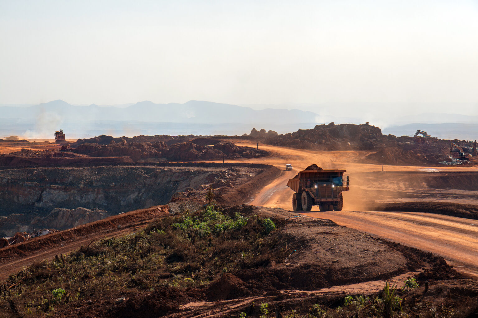 Direitos dos atingidos são o foco desta imagem, que revela uma extensa área de mineração a céu aberto, marcada por poeira, solo degradado e intensa movimentação de caminhões. O cenário evidencia os impactos sociais e ambientais que recaem sobre comunidades próximas — perda de território, contaminação da água, ruptura de modos de vida e risco à saúde. A foto reforça a urgência de garantir reparação integral, participação efetiva e justiça socioambiental, assegurando que o progresso econômico não se sobreponha aos direitos humanos.