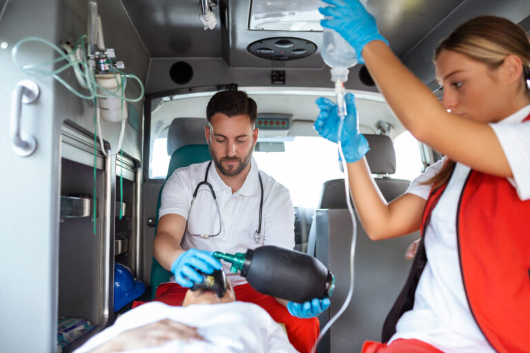 Samu: a imagem mostra o interior de uma ambulância em operação, onde dois profissionais de saúde realizam um atendimento de emergência. Um deles, usando luvas e estetoscópio, faz ventilação manual em um paciente deitado na maca, enquanto o outro ajusta um soro intravenoso. A cena retrata um momento de alta concentração e trabalho em equipe, destacando a importância do preparo técnico, da agilidade e da atuação humanizada dos socorristas. É uma imagem ideal para ilustrar temas como primeiros socorros, atendimento pré-hospitalar, serviços de emergência e a rotina dos profissionais que salvam vidas todos os dias.